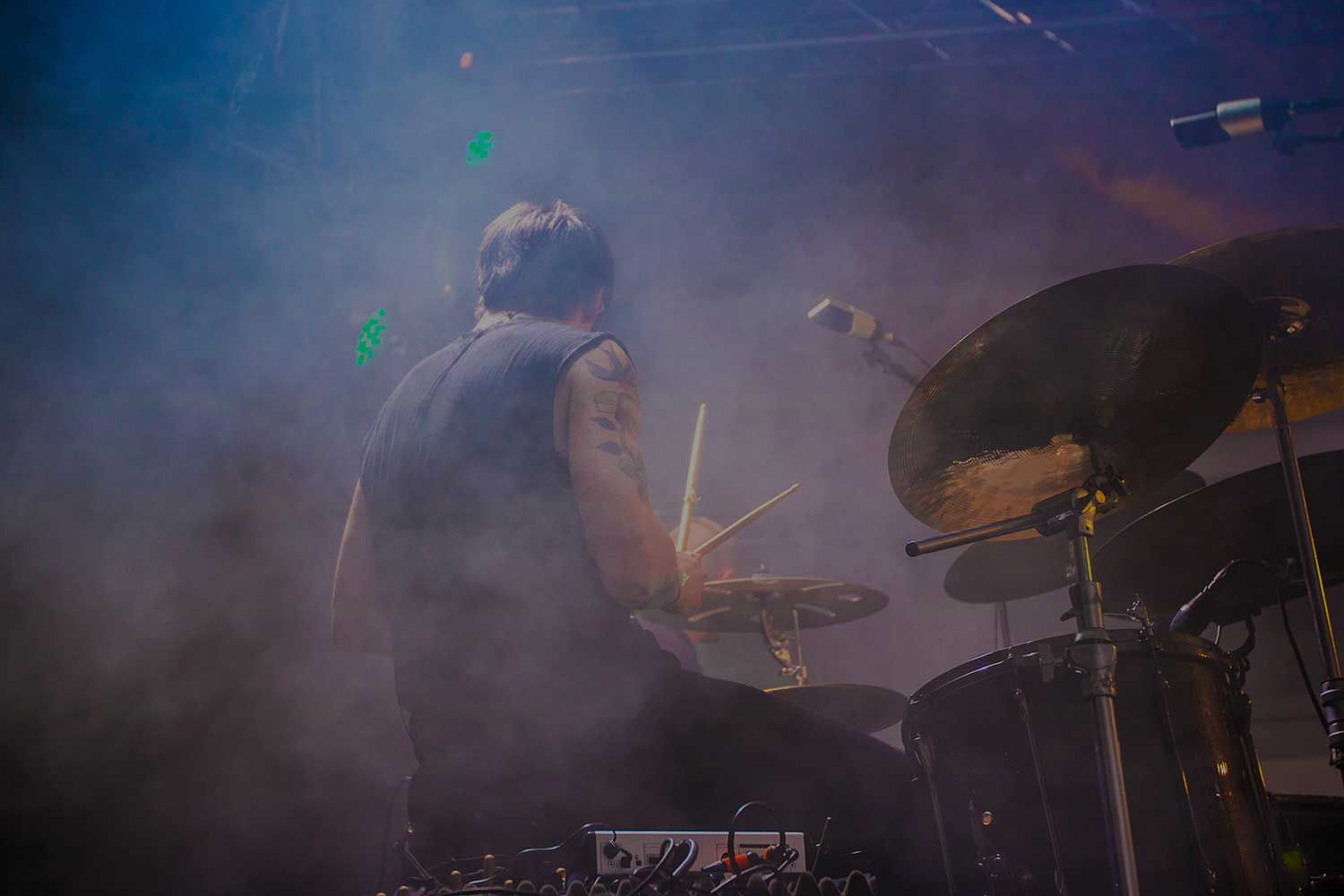 A drummer plays a drum set on a foggy stage, with various equipment and microphones surrounding him.