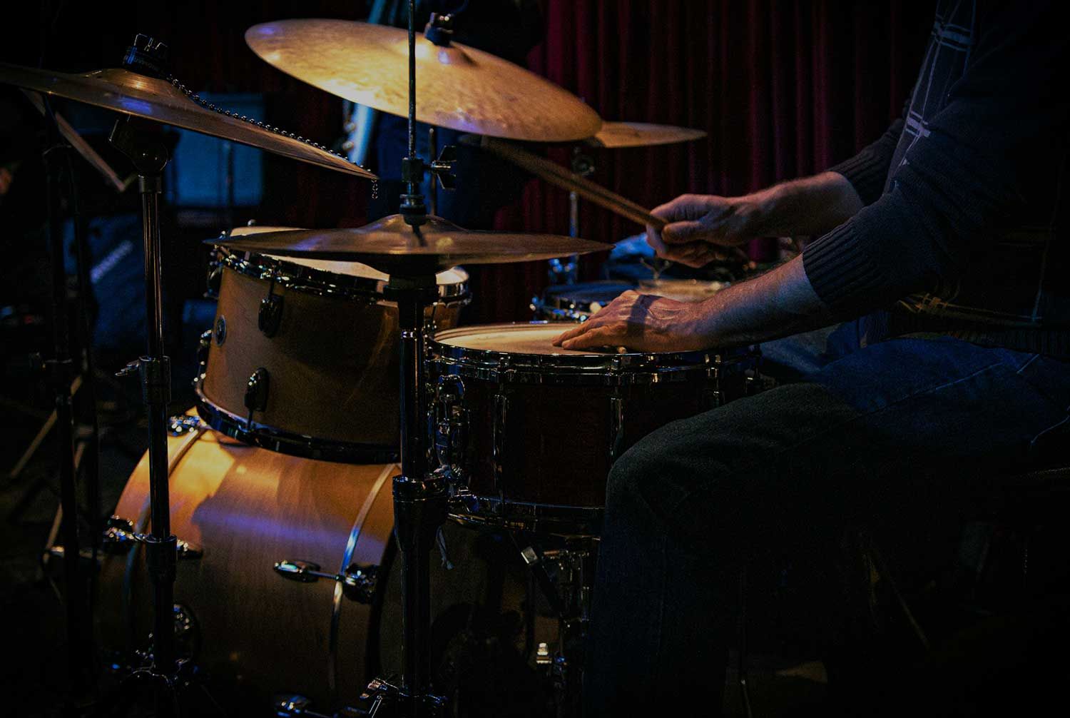 A person playing a drum set on stage, focusing on their hands and drumsticks. The background is dimly lit.