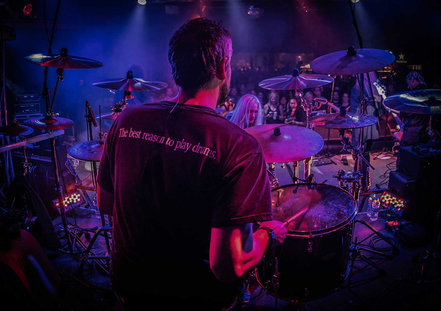 Drummer on stage playing drums with a visible t-shirt message "The best reason to play drums!" while an audience looks on from behind colorful lighting.