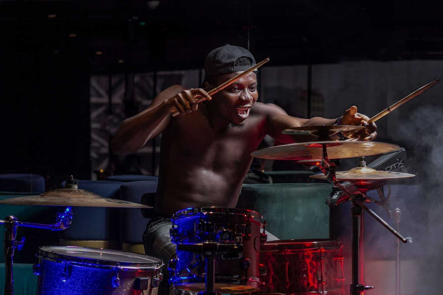 A shirtless man wearing a backward cap passionately plays the drums on a dimly lit stage.